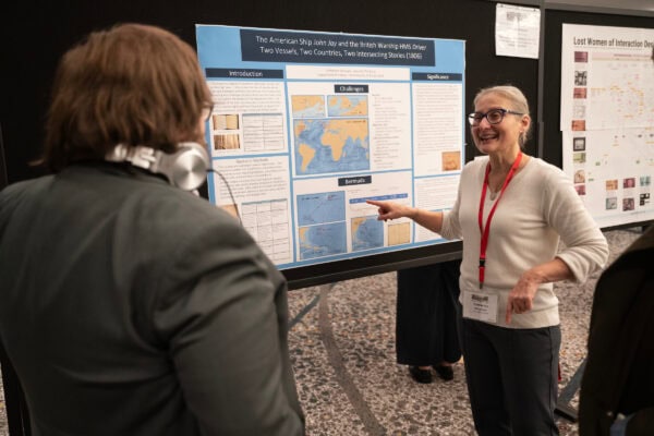A woman pointing and smiling while presenting a large poster to a man wearing headphones around his neck