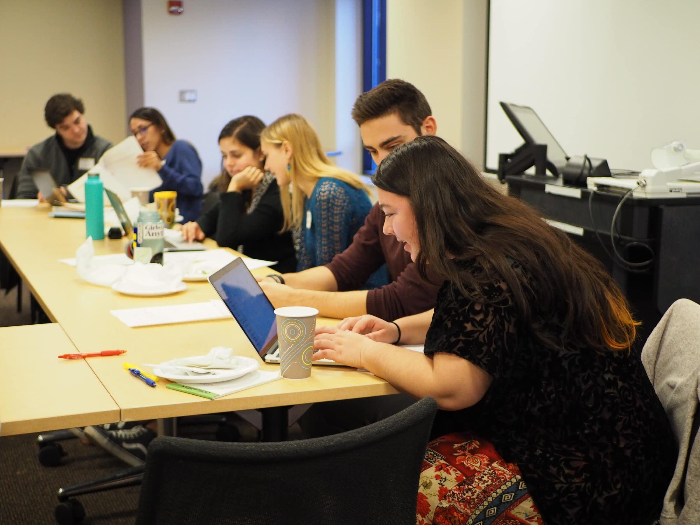 Six students sit at a long wooden table in pairs reading laptops and papers.
