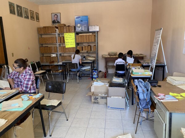 Two women sit in a small room surrounded by archival boxes, shelves, and documents.