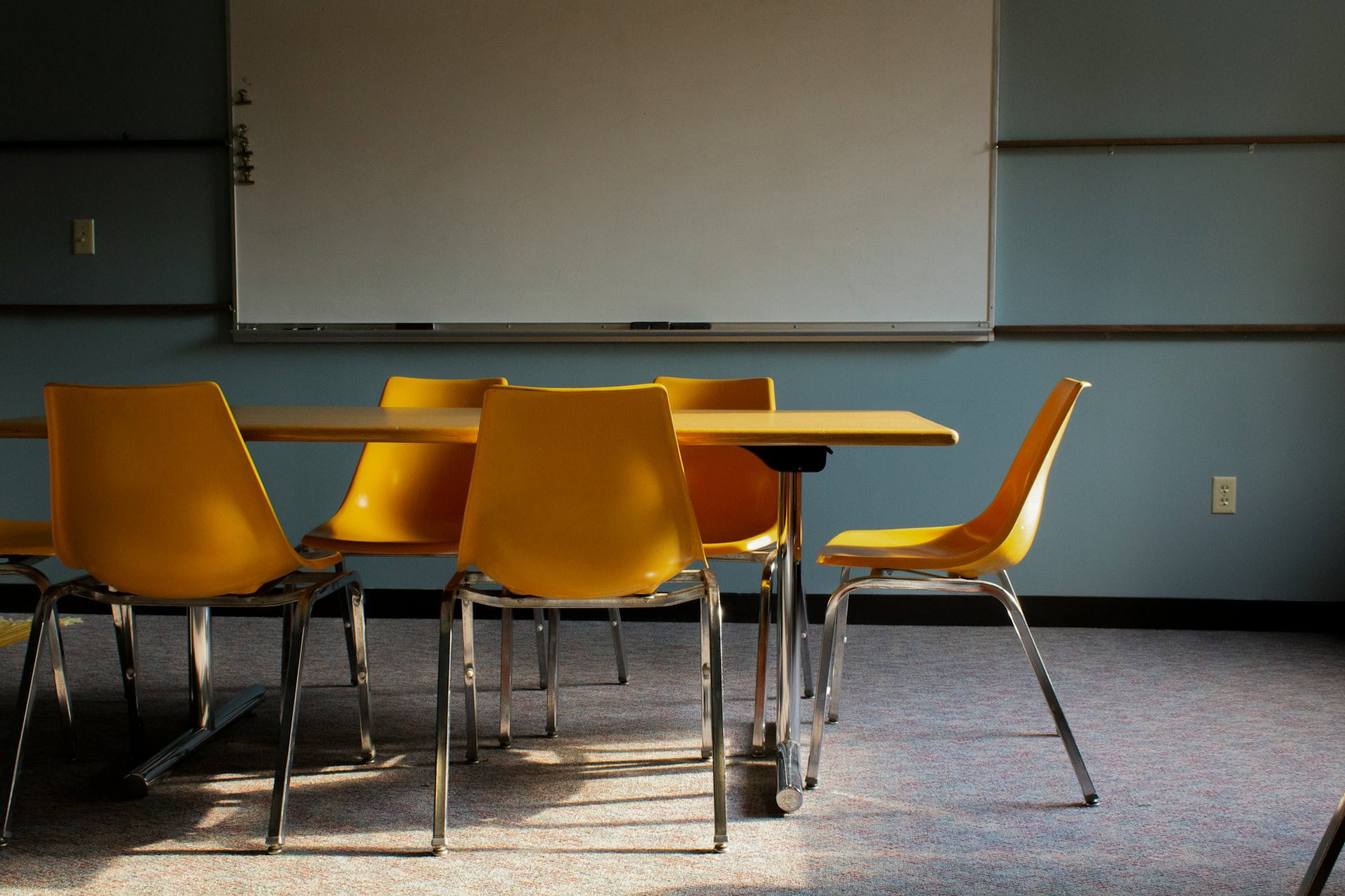 2024_Stock_YellowChairs Five yellow chairs with metal legs around a yellow chair with metal legs in a classroom in front of a whiteboard on a blue wall.