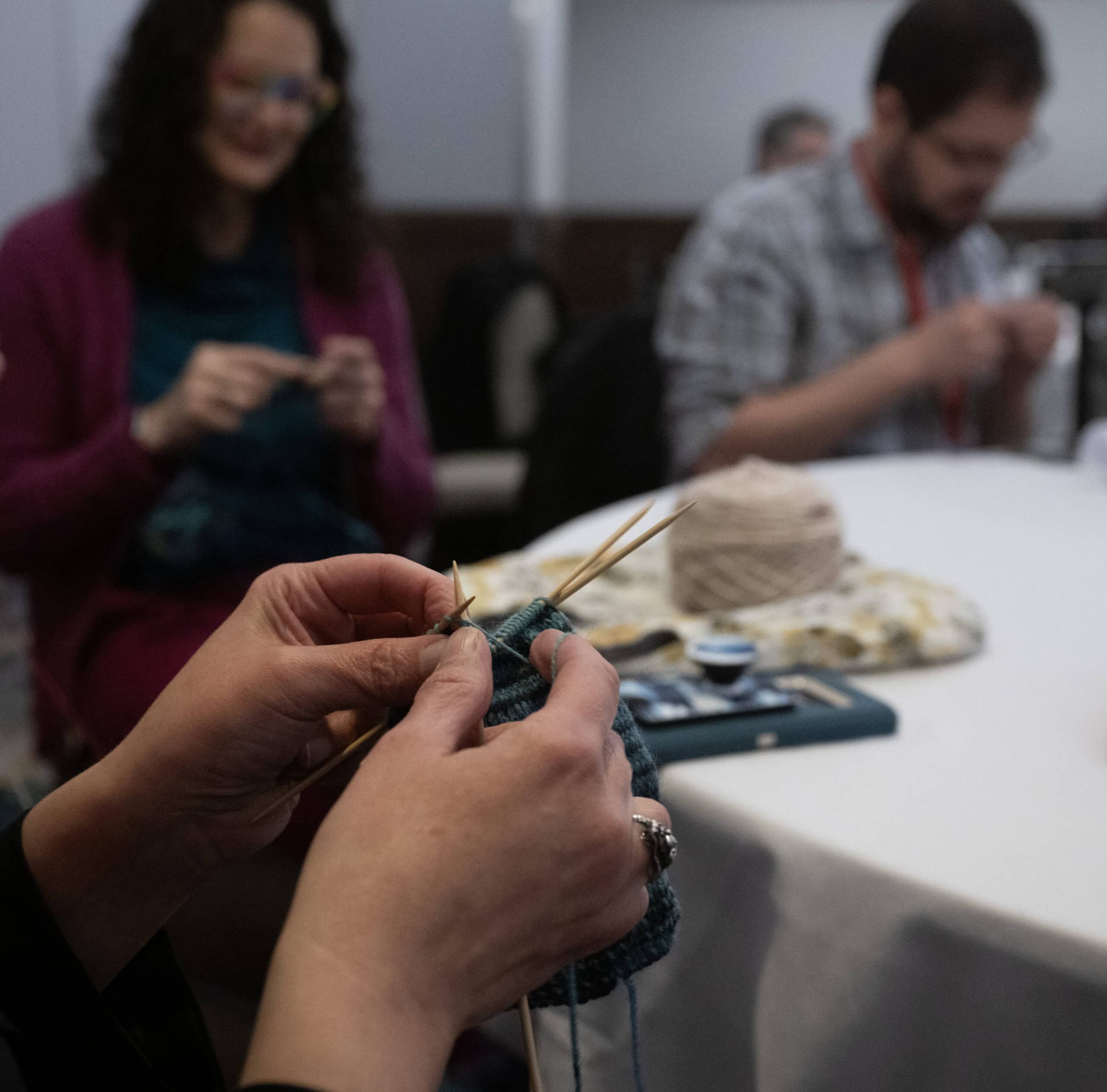 Two hands knitting with green yarn. In the background, you can see two other people working on crafts, and a table with a white tablecloth with craft supplies sitting on it.