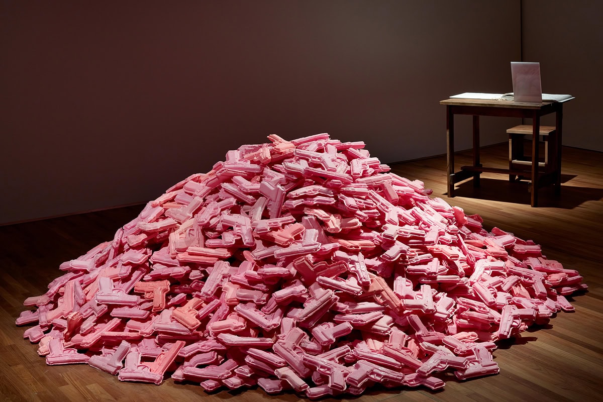 Photograph of a large pile of pink crackers in the shape of guns. In the back right corner of the exhibit, a small table and chair are visible.