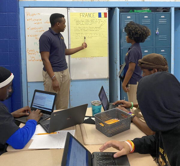 Two young people present information in front of three teens sitting at a table. The standing young man is pointing with a pen at a poster with "France" and a French flag at the top, as a young woman looks on while holding a laptop.