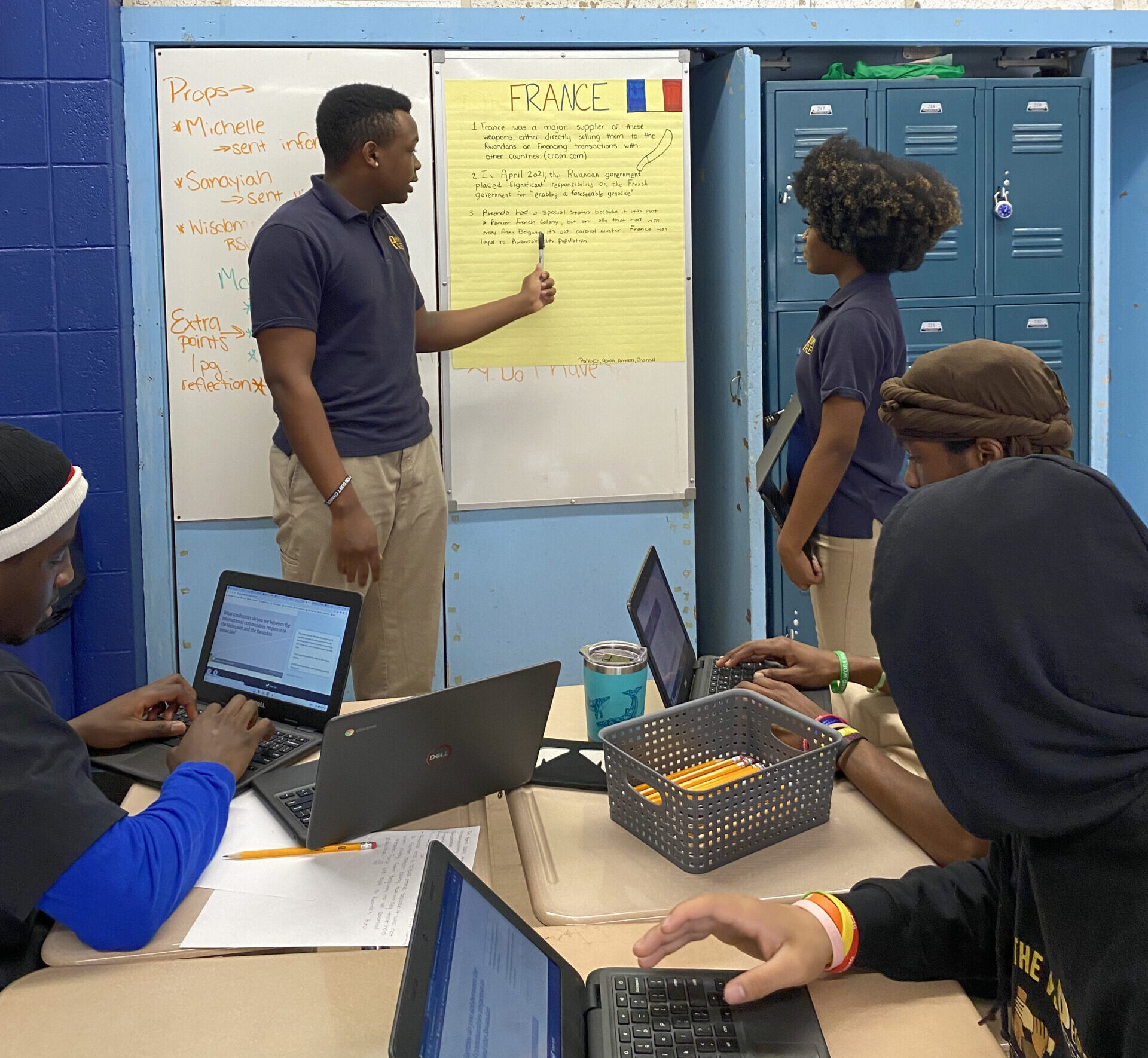 Two young people present information in front of three teens sitting at a table. The standing young man is pointing with a pen at a poster with "France" and a French flag at the top, as a young woman looks on while holding a laptop.