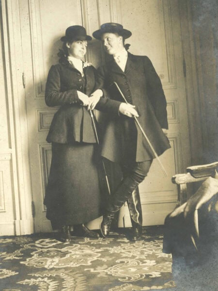 Two women leaned against a doorway looking at each other lovingly. The one on the left is wearing coat a long skirt and the one on the right is waring a coat and pants