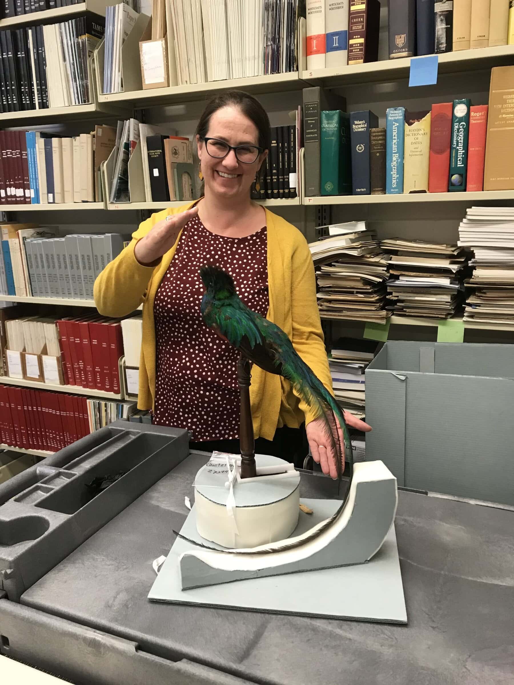 Sarah Weicksel standing behind a quetzal bird surrounded by books