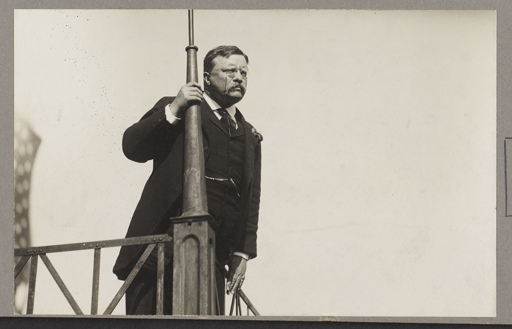 Teddy Roosevelt speaking out from a balcony holding onto a pole