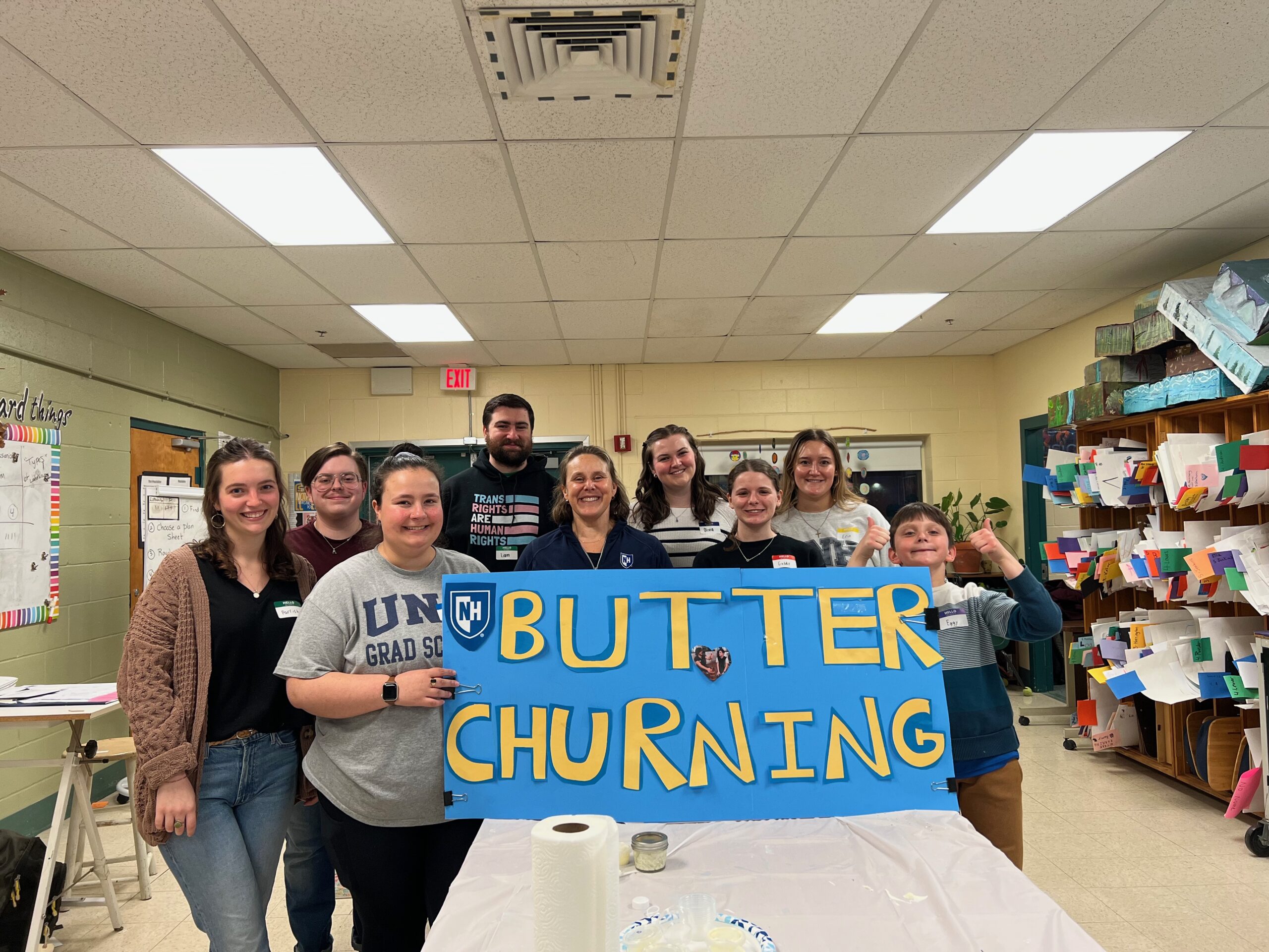 A group of adults and students holding up a painted sign that says "Butter Churning"