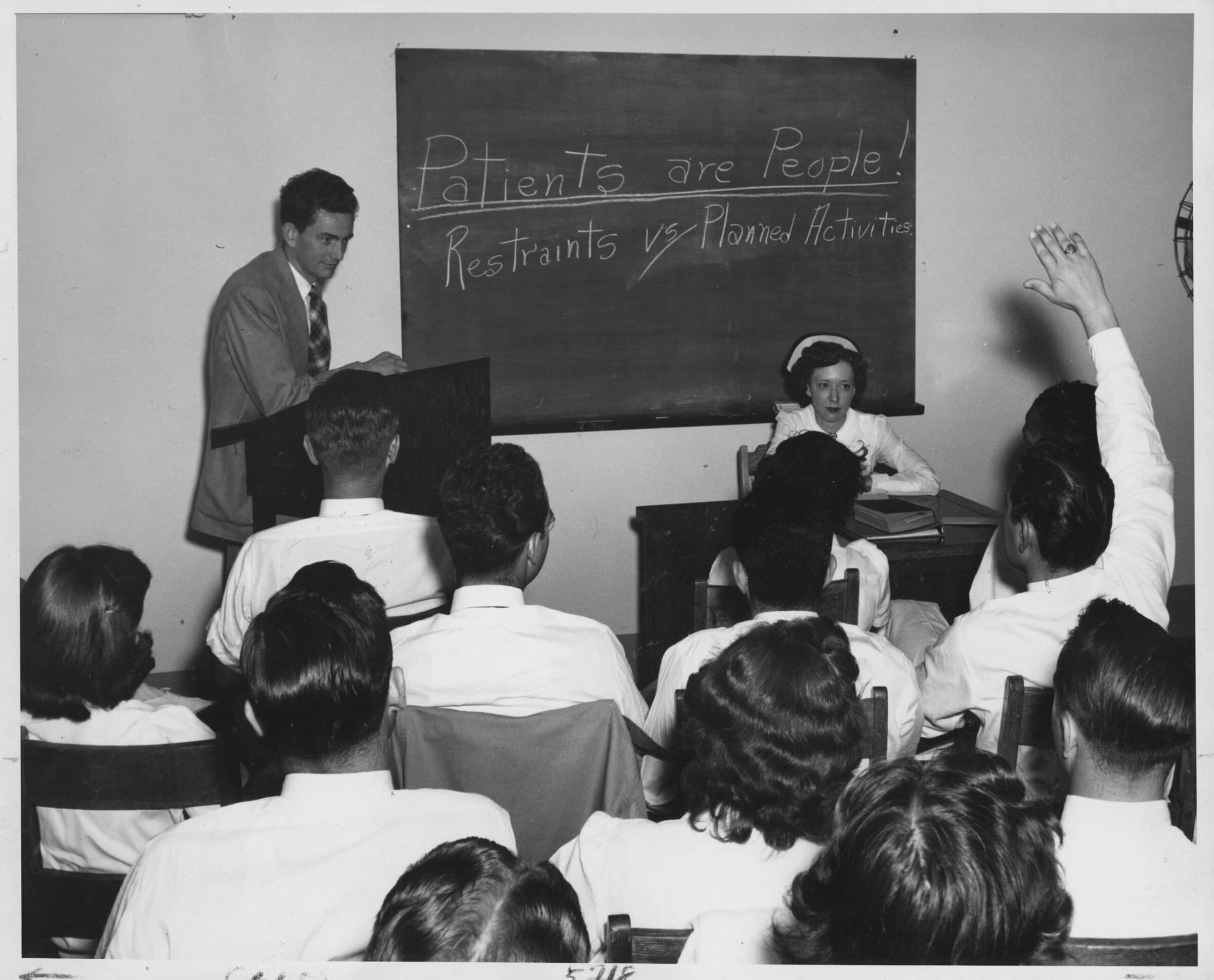 A clsasroom of nurses facing the front. On a blackboard, "Patients are People! Restraints vs Planned Activities" is written.