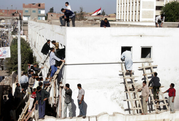 Photograph showing a white square building. On two sides, about two dozen men are waiting to climb through second story windows into the building via ladders. Several men stand on the roof, along with the Egyptian flag.
