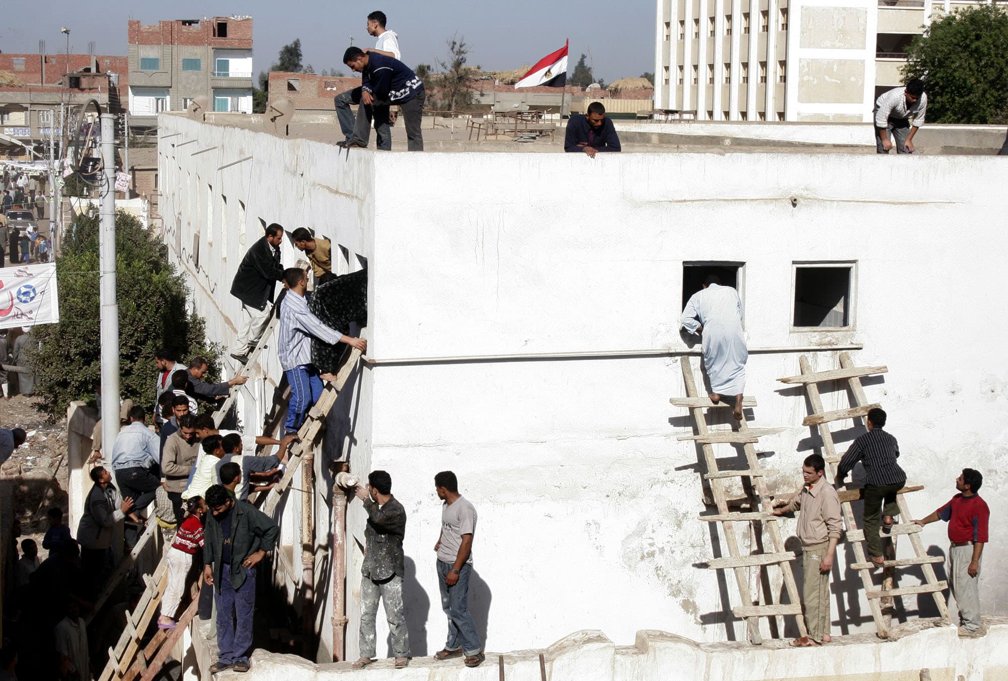 Photograph showing a white square building. On two sides, about two dozen men are waiting to climb through second story windows into the building via ladders. Several men stand on the roof, along with the Egyptian flag.