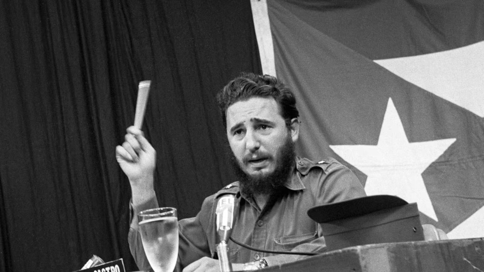 Black and white photograph of Castro sitting at a table speaking into a microphone.