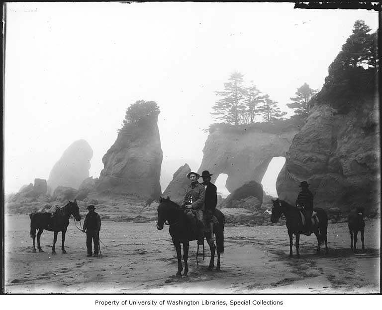 Black and white photograph of four horses with four riders along the beach. Rock structures are visible in the background.