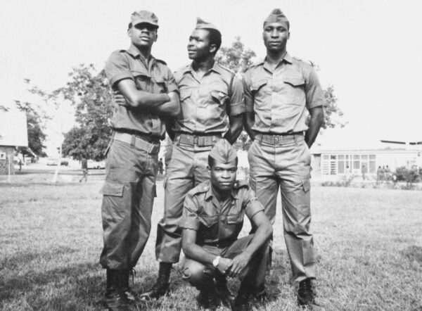 Photograph showing four young men outside in military uniforms. Three are standing in a row and one is kneeling in front of them. All are facing the camera and buildings can be seen in the background