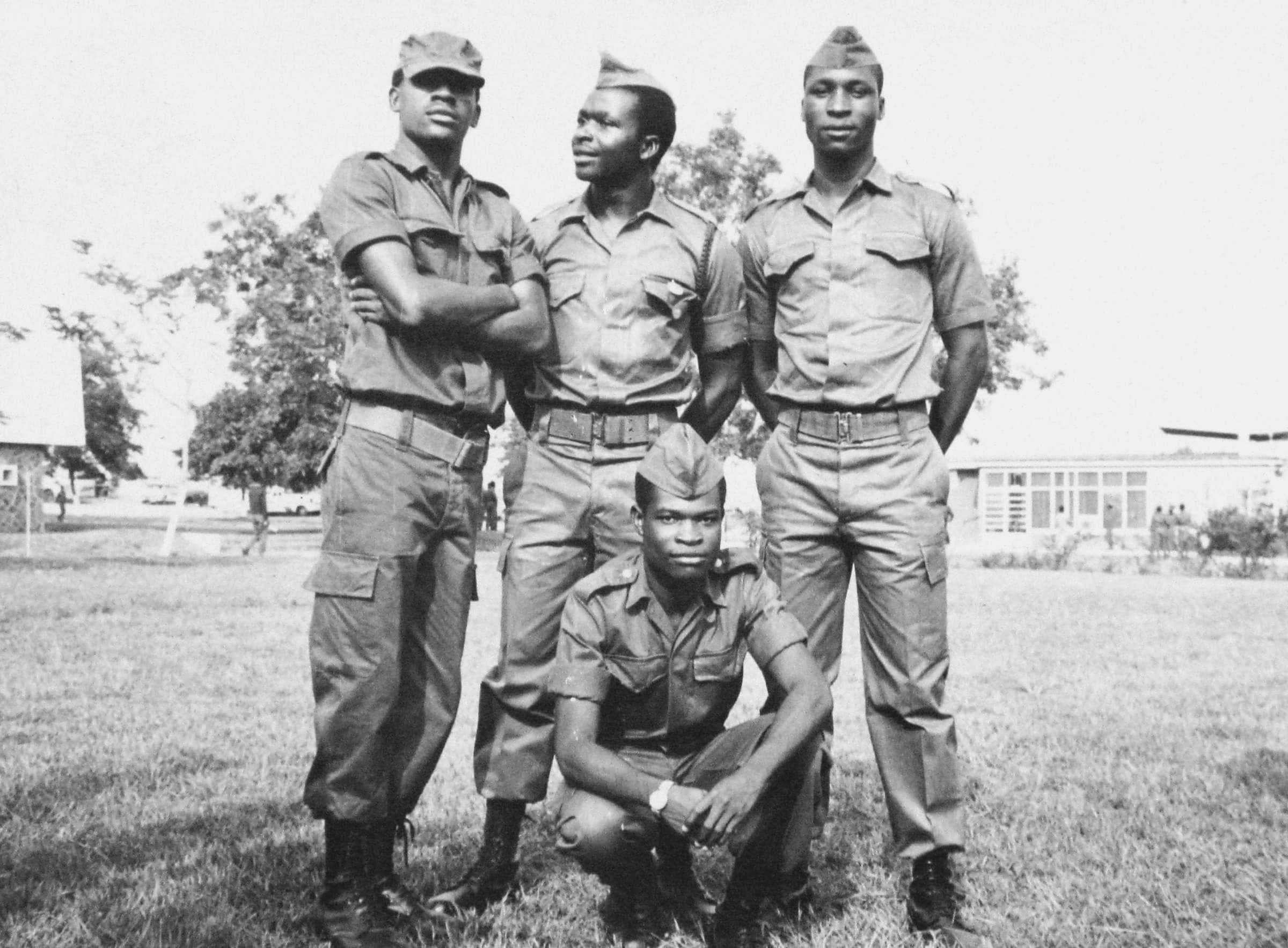 Photograph showing four young men outside in military uniforms. Three are standing in a row and one is kneeling in front of them. All are facing the camera and buildings can be seen in the background