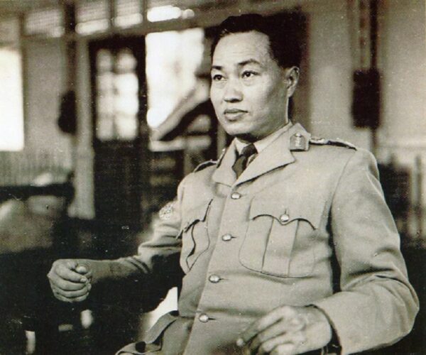 General Ne Win is shown sitting in chair looking to the side of the camera. He is wearing a formal military jacket. The background is blurred, leaving the focus on him. 