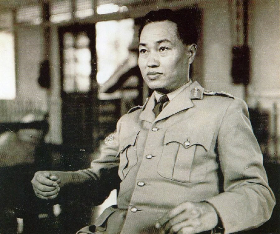 General Ne Win is shown sitting in chair looking to the side of the camera. He is wearing a formal military jacket. The background is blurred, leaving the focus on him.