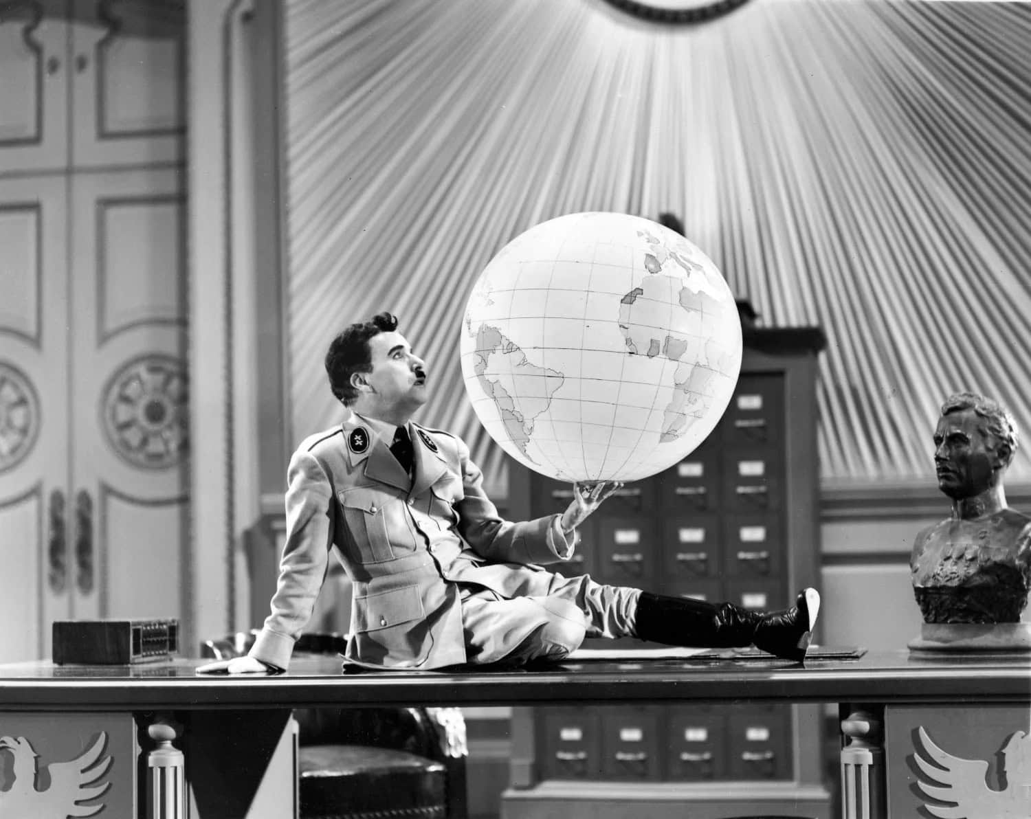 Charlie Chaplin, dressed as a Hitler-like character, sits atop a large desk. he is holding a large globe in one hand and is gazing up.