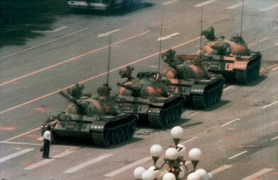Color photograph of a man standing in front of four military tanks on an empty street.