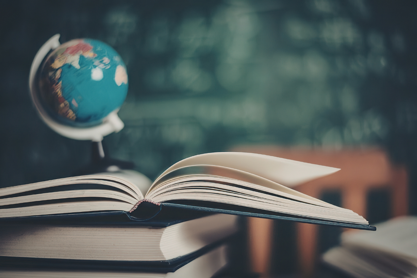 A stack of books with the top book open and a globe and chalkboard in the background.