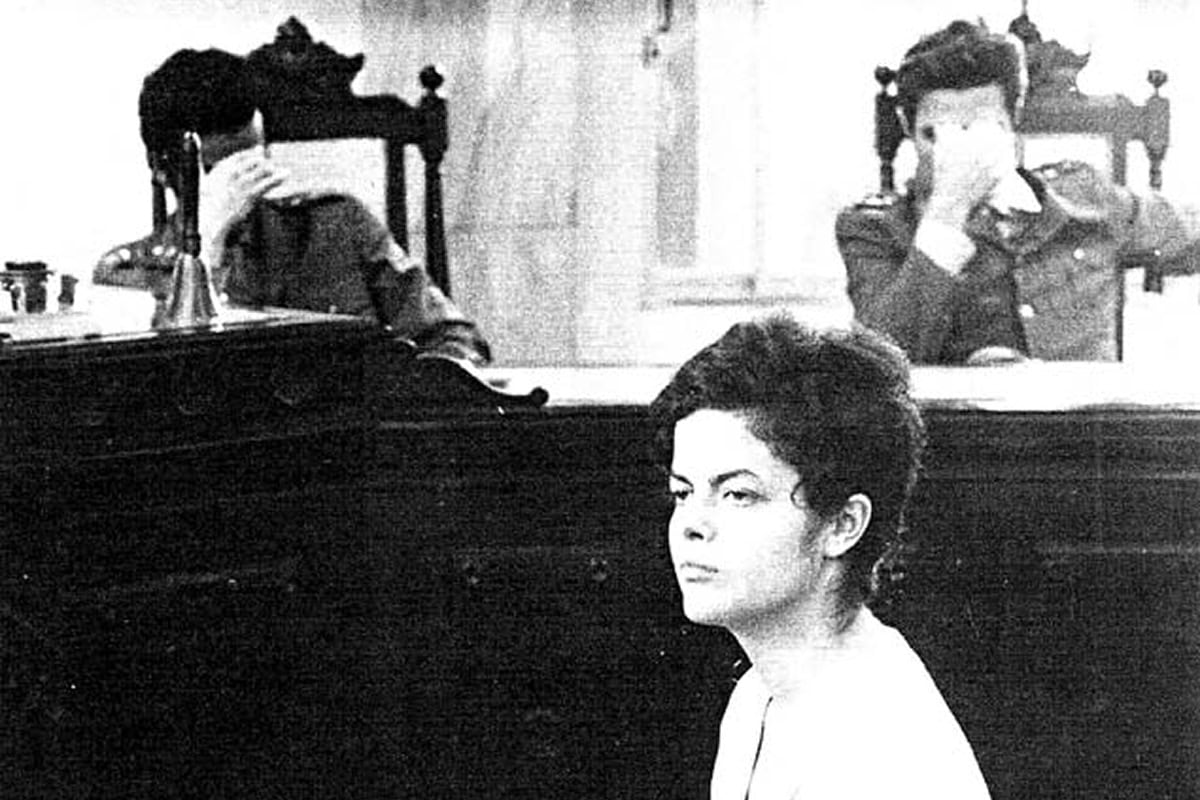 Black and white photograph of Dilma sitting with her hands folded in a chair. Two men can be seen behind her covering their faces.