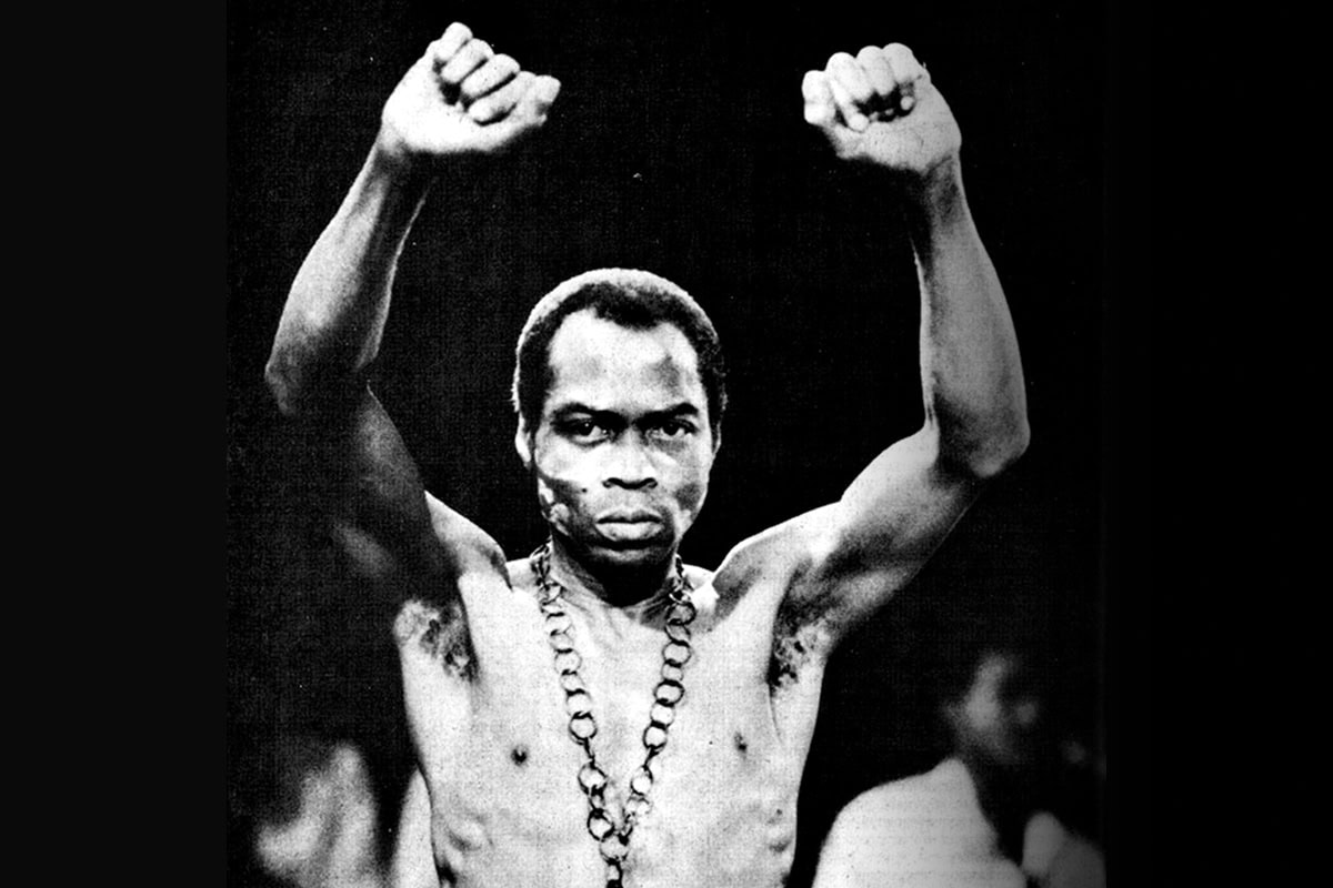 Photograph of Fela staring unsmiling at the camera. His hands are in fists and both arms are raised above him.