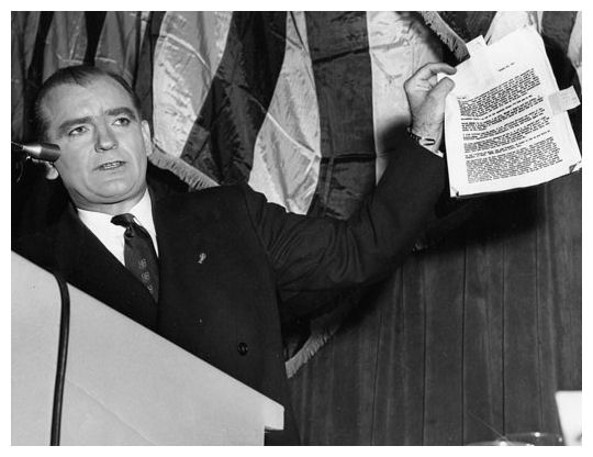 Photograph of McCarthy standing in front of a microphone, holding a document with text printed on it.