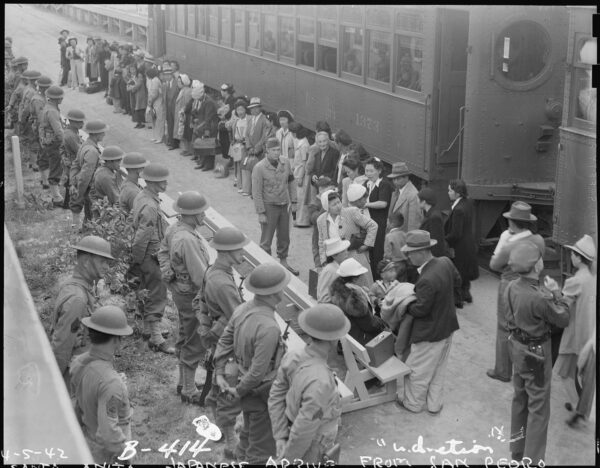Black and white photograph of a train station. A line of people wait alongside the train. Standing opposite of them is a line of men in military uniforms.