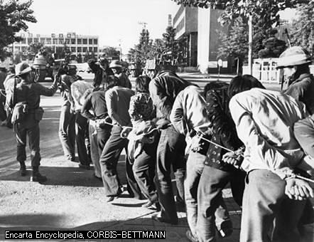 Black and white photograph of people with their hands cuffed behind their back and attached to a rope. A man in a military uniform is shown at the front of the line directing them, and another is standing alongside them. A few buildings and trees are visible int he background.