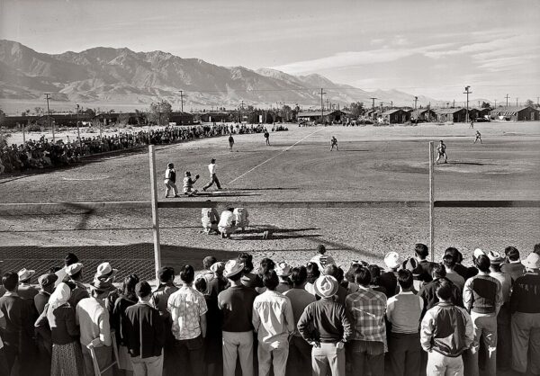 A baseball game is taking place at a dusty field in front of a crowd. Spectators are gathered closely behind a low fence. Mountains rise in the background. The scene is set in an outdoor, rural area with a clear sky.