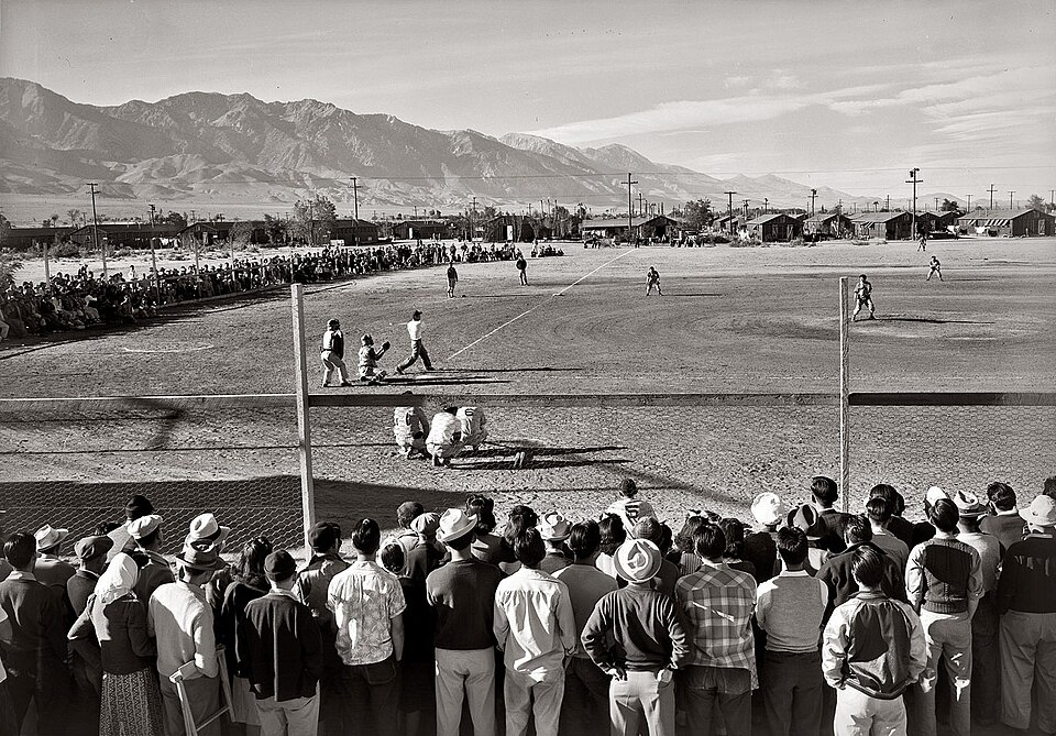 A baseball game is taking place at a dusty field in front of a crowd. Spectators are gathered closely behind a low fence. Mountains rise in the background. The scene is set in an outdoor, rural area with a clear sky.