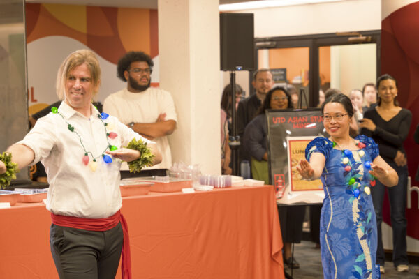 A lively scene at a cultural event featuring two people performing the hula. The person on the left wears a white top with colorful lights, a red sash, and greenery on their wrists. The person on the right wears a vibrant blue dress with floral patterns. Behind them, a group of people watches the performance near a sign that reads "UTD Asian Studies."