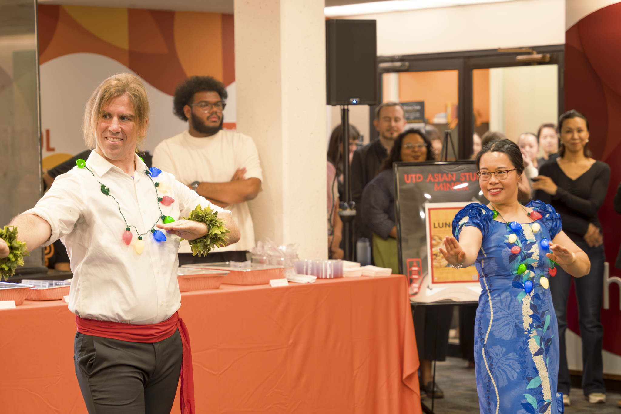 A lively scene at a cultural event featuring two people performing the hula. The person on the left wears a white top with colorful lights, a red sash, and greenery on their wrists. The person on the right wears a vibrant blue dress with floral patterns. Behind them, a group of people watches the performance near a sign that reads "UTD Asian Studies."