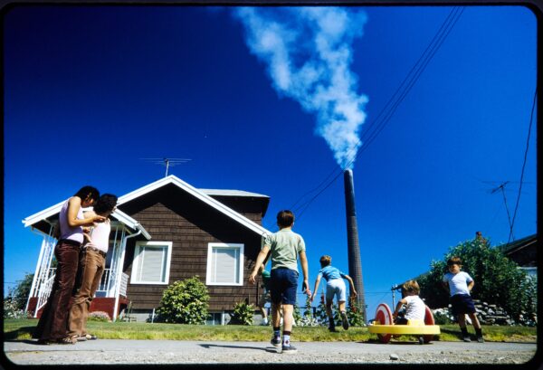 A group of people are in front of a house with a brown exterior. A large smokestack in the background emits a plume of smoke under a clear blue sky. Children are playing with toys on the sidewalk.