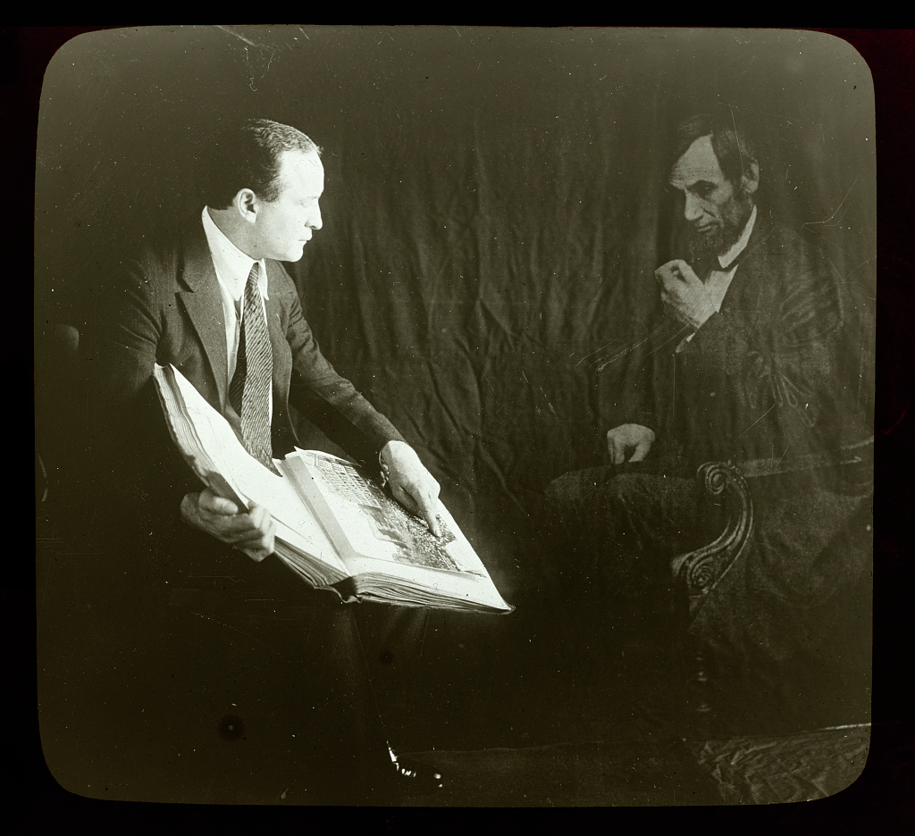 A person is sitting in a chair, holding an open book and facing a transparent figure of Abraham Lincoln. The background is dark, creating a ghostly effect for Lincoln's figure.
