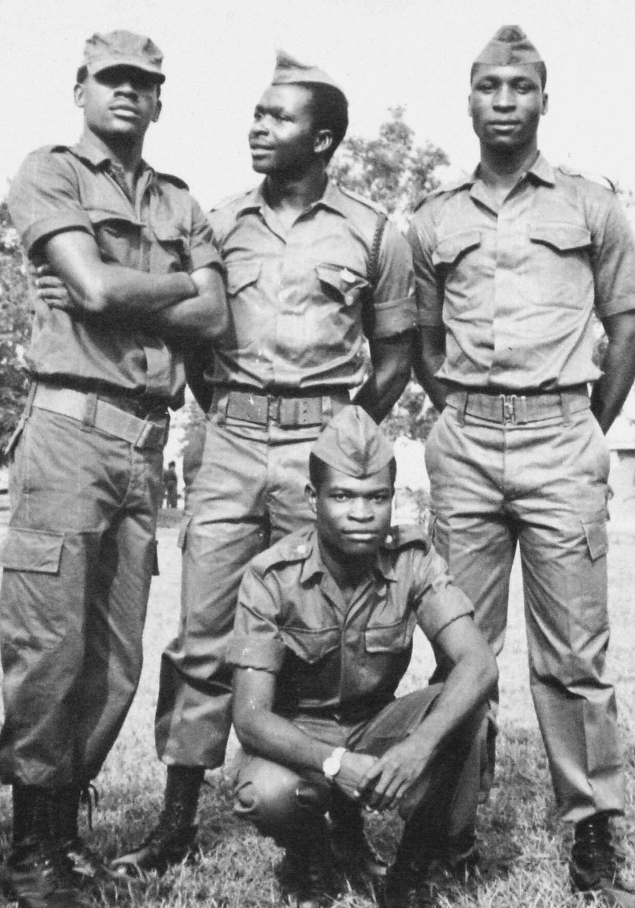Photograph showing four young men outside in military uniforms. Three are standing in a row and one is kneeling in front of them. All are facing the camera and buildings can be seen in the background.